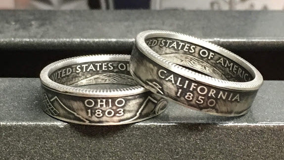 Two silver coin rings with 'United States of America' and state names engraved on them, placed on a dark surface.
